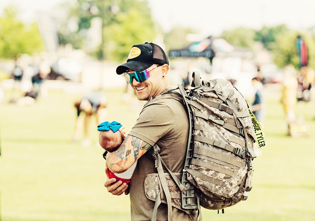 Veteran in Bass Pro Shop hat, glasses and backpack holding a small baby