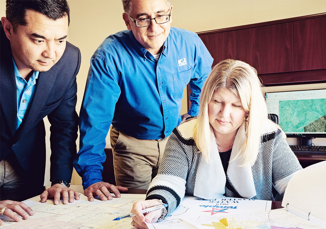 Two men oversee a woman as she shows them maps