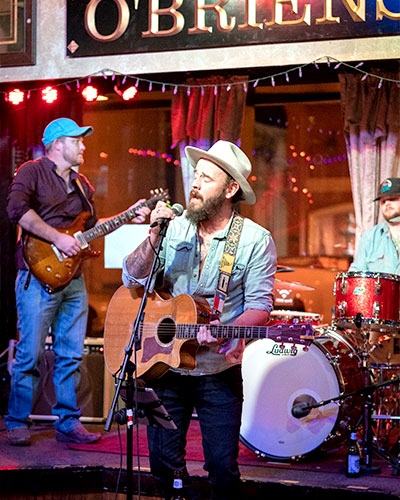 Man in a hipster outfit holding an acoustic guitar sings passionately into a mic