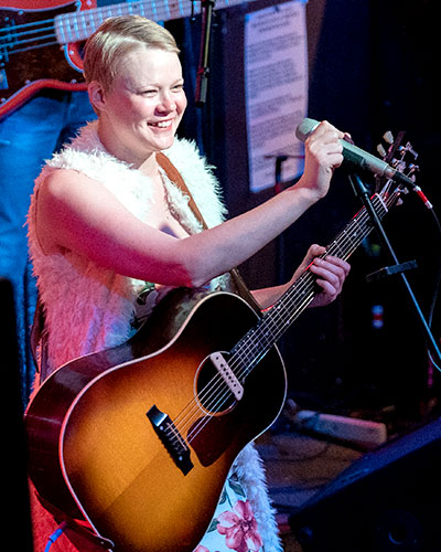 Female singer holding an acoustic guitar grabs the microphone while smiling at the audience