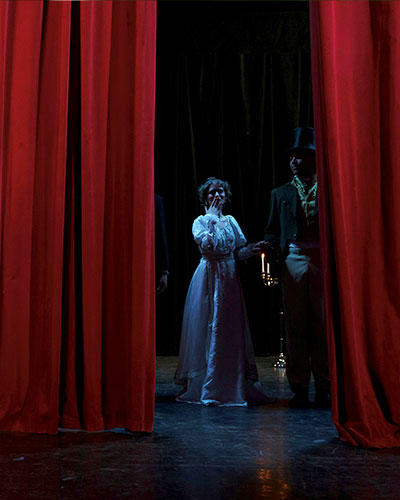 Two thespians in Victorian garb looking at the camera from behind a red curtain on a stage