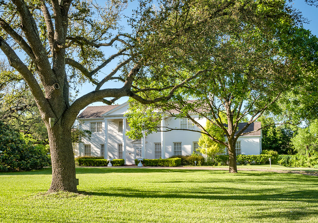 Large whtie plantation-style home behind a yard with two trees