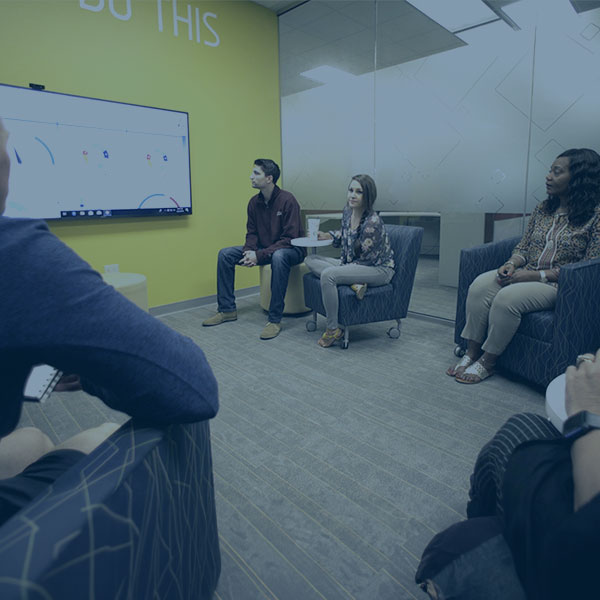 People in a small meeting room sitting in couches looking at a screen and each other