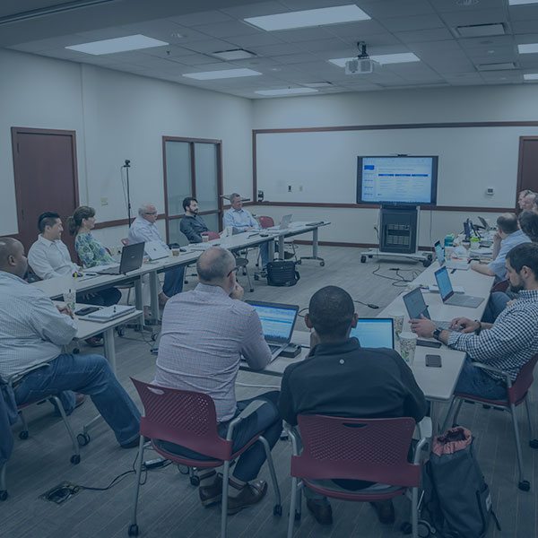 Group of people sitting in front of laptops in a semi-circle formation ina conference room looking at a screen