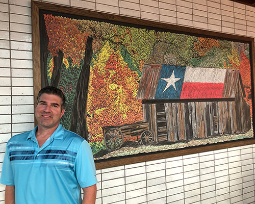 Man stands in front of a painting of a cabin with the Texas flag painted on the roof