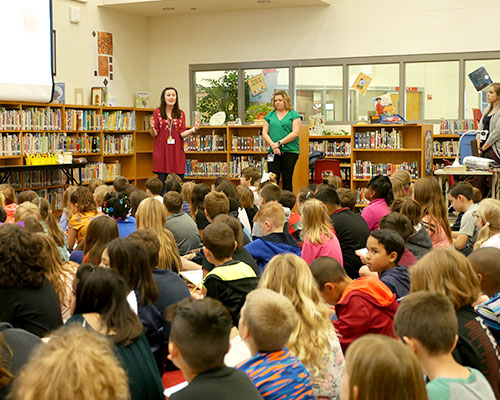 Two teachers instruct a library full of children sitting cross-legged