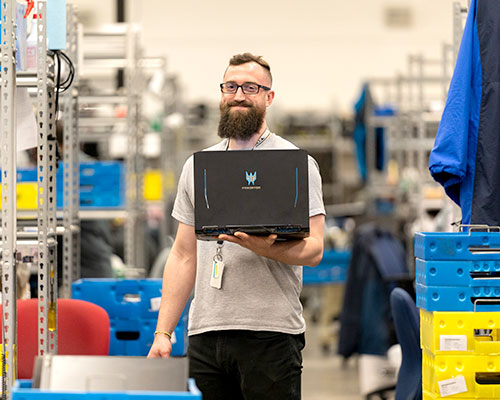 Man with bushy beard in a warehouse holding a black laptop while smiling at camera