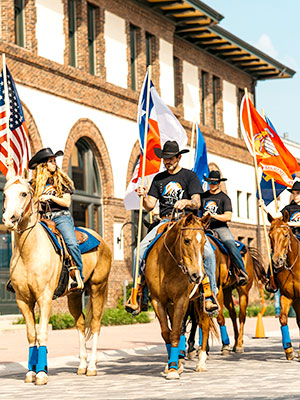 People with cowboy hats holding flags riding on horses walking next to a building