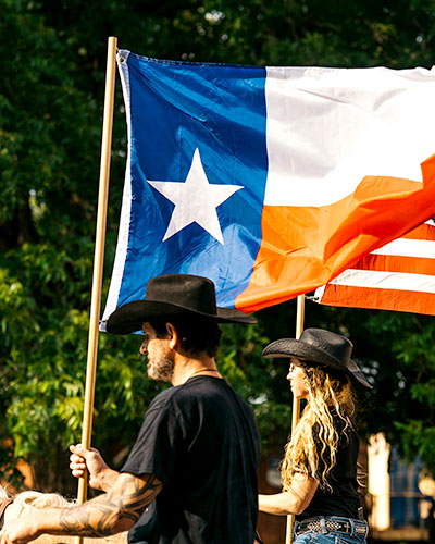 A man and woman in cowboy hats ride horses while holding the Texas and American flag