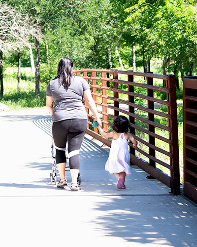 Woman walks hand-in-hand with her daughter on a paved path through a park