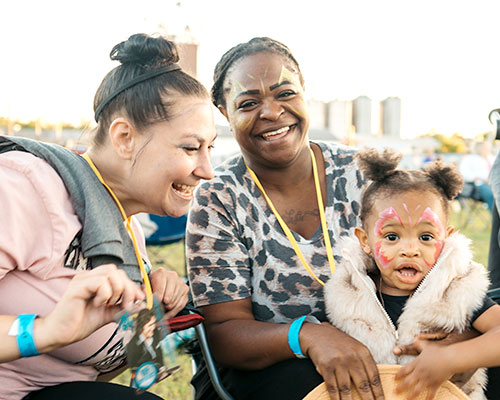 Two women and young toddler with a butterfly pattern on face smile towards camera